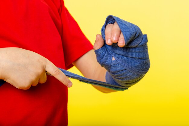 Ein kleiner Kindjunge der Vorderansicht, der seine Hand mit blauem Taschentuch für das Boxen im roten T-Shirt an der gelben Wand bindet