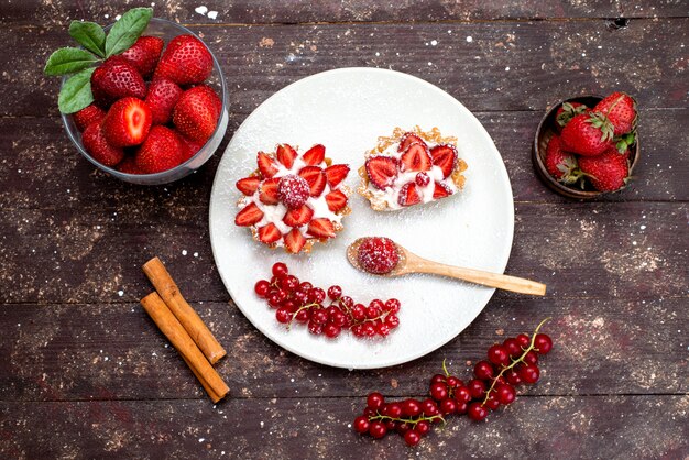 Ein kleiner Kuchen von oben mit Sahne und geschnittenen frischen Erdbeeren in weißer Platte zusammen mit Zimt auf dem braunen Schreibtischobstkuchen