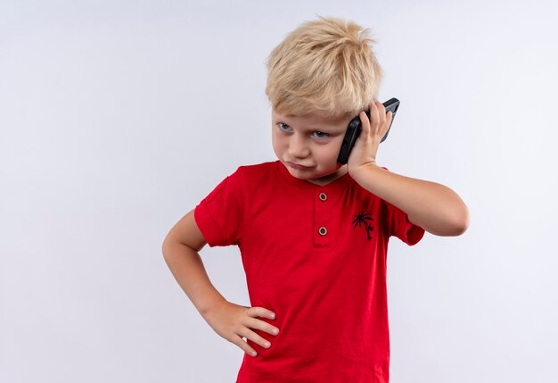 Ein kleiner süßer blonder Junge im roten T-Shirt spricht auf Handy mit Hand auf Taille auf einer weißen Wand