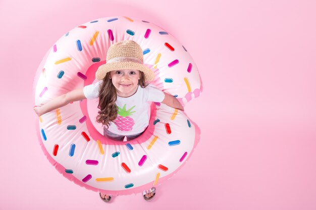 Ein kleines Kindermädchen in Freizeitkleidung, das auf einem aufblasbaren Donutkreis liegt. Rosa Hintergrund. Draufsicht. Sommerkonzept.