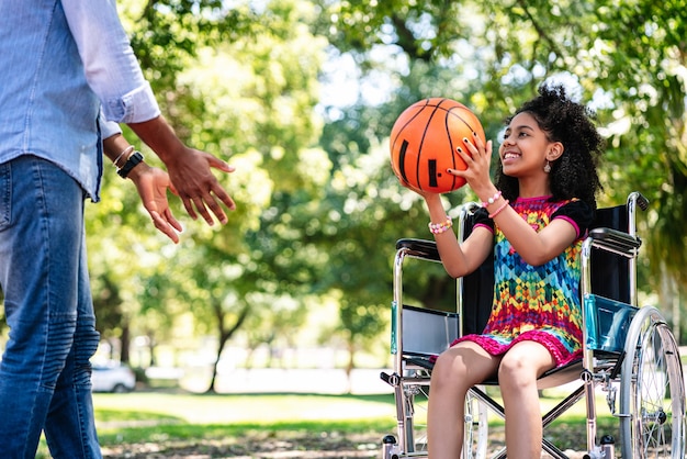Ein kleines Mädchen im Rollstuhl, das Spaß mit ihrem Vater beim gemeinsamen Basketballspielen im Park hat