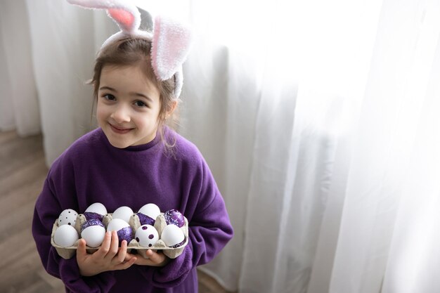 Ein kleines Mädchen mit Osterohren hält ein Tablett mit Feiertagseiern