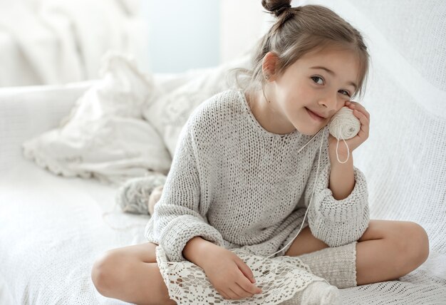 Ein kleines Mädchen sitzt mit einem Fadenknäuel auf dem Sofa und lernt stricken.