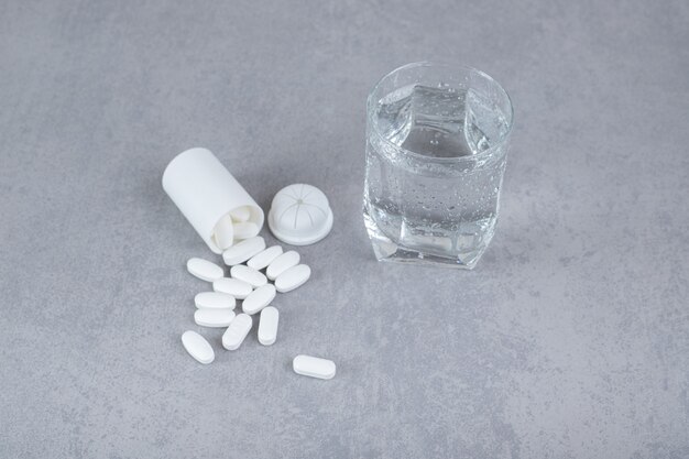 Ein kleines weißes Glas mit weißen Pillen mit einem Glas reinem Wasser auf grauer Oberfläche