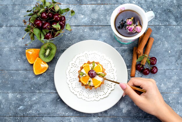 Ein köstlicher kleiner Kuchen von oben mit Sahne und frisch geschnittenen Früchten zusammen mit Zimttee auf dem grau-blauen Schreibtischfruchtkuchen-Kekstee