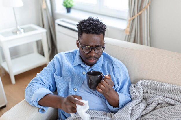 Ein kranker afrikanischer junger Mann, der mit einer Decke bedeckt ist, die eine laufende Nase bläst, hat Fieber erwischt, das beim Niesen in Gewebe erkältet ist, sitzt auf dem Sofa, ein kranker allergischer schwarzer Mann mit Allergiesymptomen, die zu Hause husten Corona 19