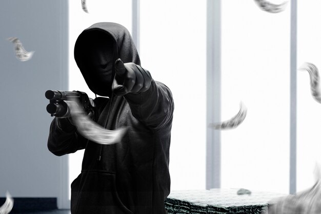 Ein krimineller Mann in einer versteckten Maske hält die Schrotflinte und zeigt auf etwas, während er das Geld auf der Bank raubt