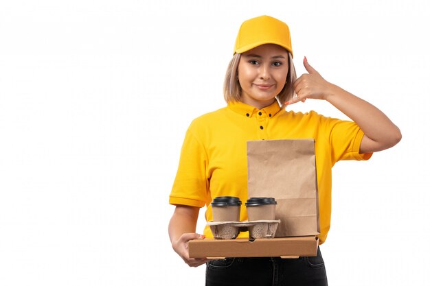 Ein Kurier der Vorderansicht weiblicher in gelber Kappe des gelben Hemdes und in den schwarzen Jeans, die das Halten des Kaffee- und Nahrungsmittelpakets aufwerfen