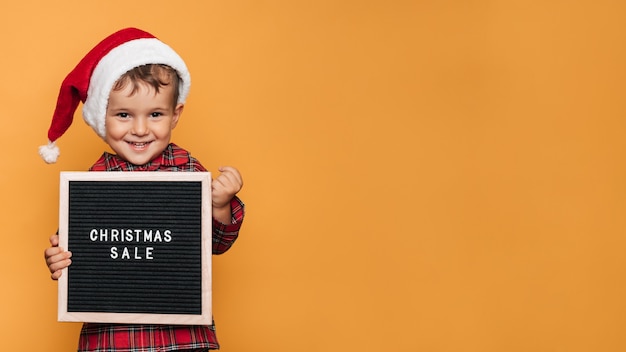 Ein lächelnder junge in einer roten weihnachtsmütze und einem pyjama, mit einem brett mit weißen buchstaben und der aufschrift frohe weihnachten in seinen händen. ein platz für ihren text und ihre werbung.