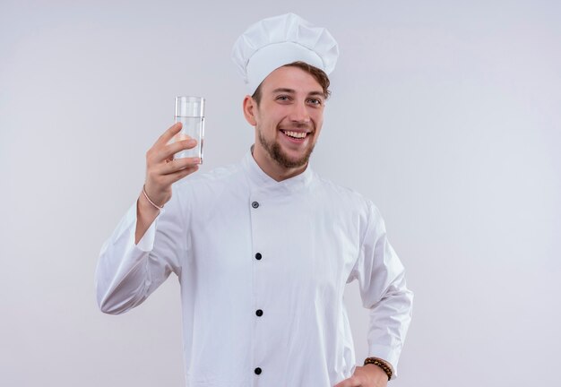 Ein lächelnder junger bärtiger Kochmann, der weiße Kochuniform und einen Hut trägt, der ein Glas Wasser zeigt, während er auf eine weiße Wand schaut