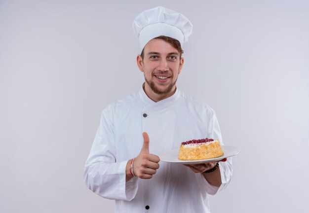 Ein lächelnder junger bärtiger Kochmann, der weiße Kochuniform und Hut trägt, hält einen Teller mit Kuchen und zeigt Daumen hoch, während er auf eine weiße Wand schaut