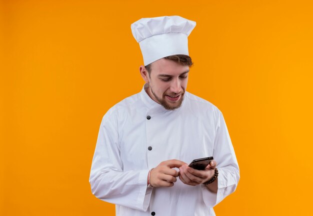 Ein lächelnder junger bärtiger Kochmann in der weißen Uniform, die sein Handy an einer orange Wand berührt