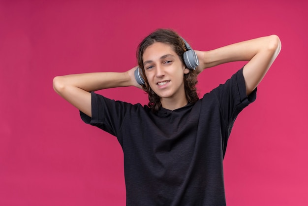 Ein lächelnder kerl mit langen haaren im schwarzen t-shirt hört musik vom kopfhörer und packt seinen kopf an der rosa wand