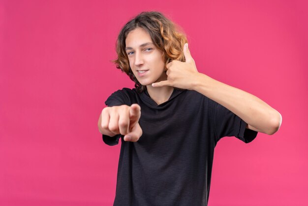 Ein lächelnder Kerl mit langen Haaren im schwarzen T-Shirt ruft mit seiner Hand an und zeigt auf die rosa Wand nach vorne