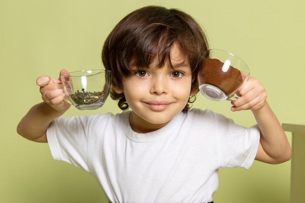 Ein lächelnder Kinderjunge der Vorderansicht, der Kaffee und Spezies im weißen T-Shirt auf dem steinfarbenen Boden hält