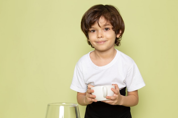 Ein lächelnder Kinderjunge der Vorderansicht im weißen T-Shirt, das Kaffeegetränk auf dem steinfarbenen Raum vorbereitet