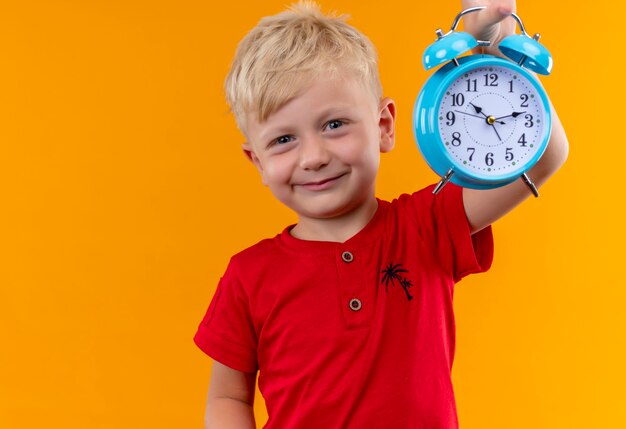 Ein lächelnder kleiner Junge mit blonden Haaren und blauen Augen, die rotes T-Shirt tragen, das blauen Wecker zeigt, während auf einer gelben Wand schaut