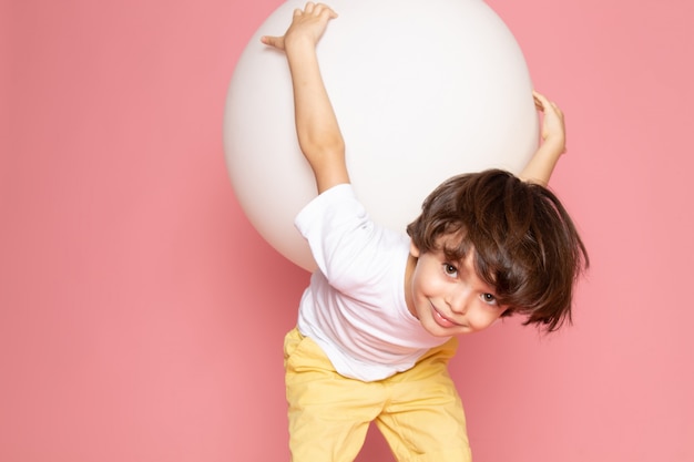 Ein lächelnder süßer Junge der Vorderansicht im weißen T-Shirt, der weißen Ball auf dem rosa Raum hält