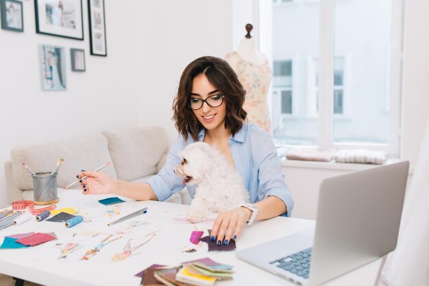 Ein lächelndes brünettes Mädchen in einem blauen Hemd sitzt am Tisch im Werkstattstudio. Sie arbeitet mit kreativen Sachen. Sie hat einen netten Hund auf den Knien.