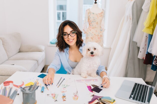 Ein lächelndes brünettes Mädchen in einem blauen Hemd sitzt am Tisch im Werkstattstudio. Sie arbeitet mit Skizzen und Stoffmustern. Sie hat einen schönen Hund auf den Knien.