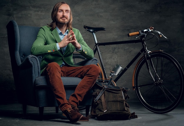 Ein lässiger bärtiger Hipster-Mann in einer grünen Jacke sitzt auf einem Stuhl mit Single-Speed-Fahrrad im Hintergrund.