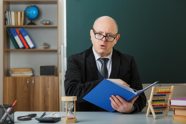 Ein Lehrer mit Brille, der das Klassenbuch überprüft und in die Kamera schaut, ist verwirrt und überrascht, wie er an der Schulbank vor der Tafel im Klassenzimmer sitzt