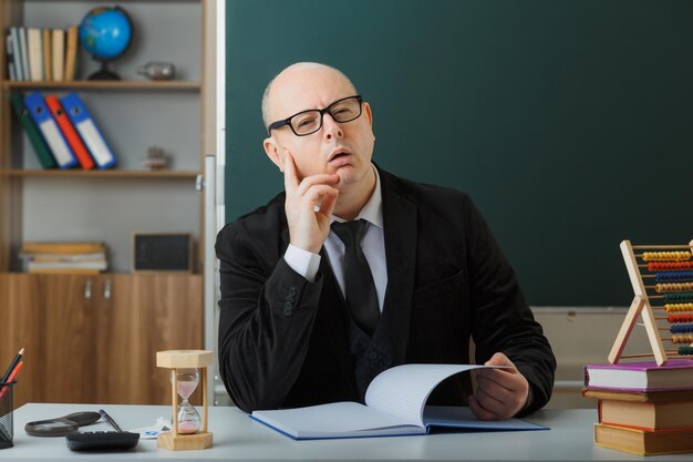 Ein Lehrer mit Brille, der das Klassenregister überprüft und verwirrt mit nachdenklichem Gesichtsausdruck sitzt, sitzt an der Schulbank vor der Tafel im Klassenzimmer