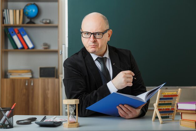 Ein Lehrer mit Brille sitzt an der Schulbank und überprüft das Klassenregister vor der Tafel im Klassenzimmer und blickt mit wütendem Gesicht in die Kamera