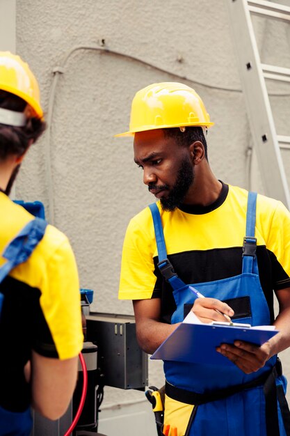 Ein lizenzierter Servicetechniker beginnt mit der routinemäßigen Wartung des Kondensators und liest mithilfe von Verteilermessgeräten den Druck in der externen Klimaanlage ab, während ein erfahrener Installateur den Prüfbericht des HVAC-Systems in die Zwischenablage schreibt