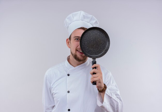 Ein lustiger junger bärtiger Kochmann, der weiße Kochuniform und Hut hält, der Bratpfanne vor seinem Gesicht hält, während auf einer weißen Wand schaut