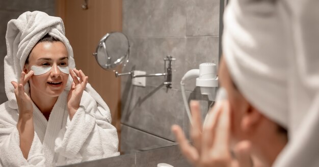 Ein Mädchen im Bademantel und mit einem Handtuch auf dem Kopf klebt im Badezimmer vor dem Spiegel Flecken unter die Augen.