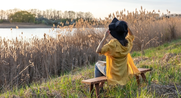 Ein Mädchen mit Hut und im lässigen Stil sitzt bei Sonnenuntergang auf einer Bank in der Nähe des Sees.