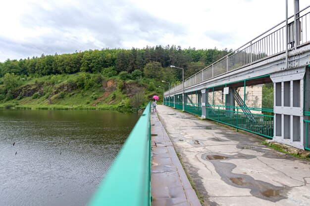 Ein Mädchen mit Regenschirm bei bewölktem Wetter für einen Waldspaziergang steht auf einer Brücke vor dem Hintergrund einer Landschaft