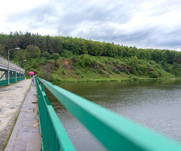 Ein Mädchen mit Regenschirm bei bewölktem Wetter für einen Waldspaziergang steht auf einer Brücke vor dem Hintergrund einer Landschaft