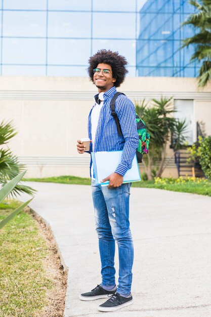 Ein männlicher Afro-Student, der Bücher und die Wegwerfkaffeetasse steht am Campus hält