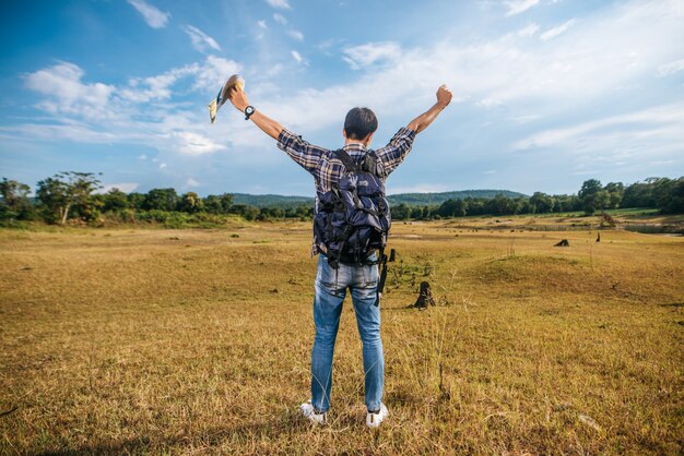 Ein männlicher Reisender mit einem Rucksack, der eine Karte trägt und auf der Wiese steht.