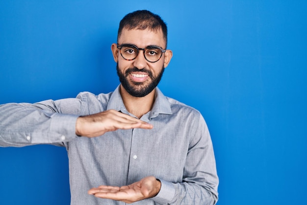 Ein Mann aus dem Nahen Osten mit Bart, der über einem blauen Hintergrund steht und mit den Händen Geste macht, die ein großes und großes Zeichen zeigen, ein Messsymbol, das lächelt und auf die Kamera schaut, das Messkonzept