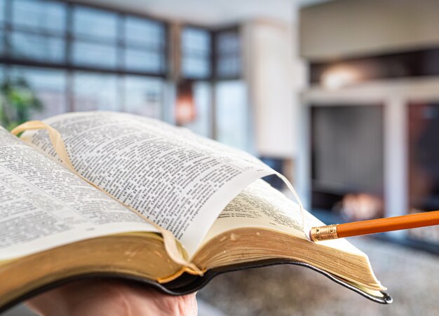 Ein Mann hält eine Bibel mit einem Bleistift vor dem Hintergrund des Wohnzimmers. In gemütlicher Atmosphäre ein Buch lesen. Nahaufnahme.