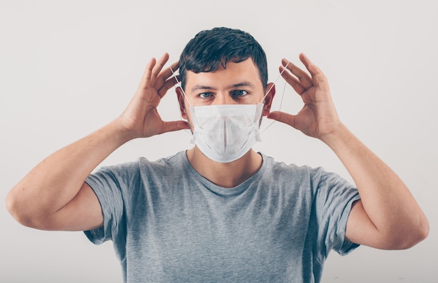 Ein Mann im grauen T-Shirt, das medizinische Maske im weißen Hintergrund trägt.