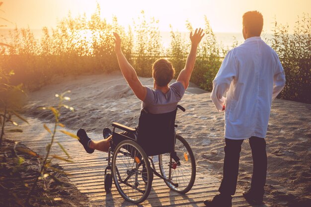 Ein Mann im Rollstuhl und seine Krankenschwester genießen den Sonnenaufgang am Strand.