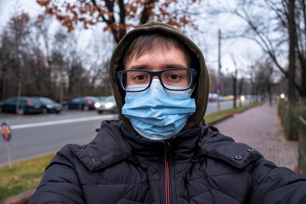 Ein Mann in Brille, medizinischer Maske, Kapuze und Jacke bei bewölktem Wetter, Blick in die Kamera, Straße auf dem Hintergrund