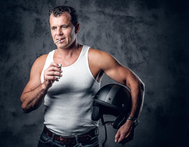 Ein Mann in einem weißen T-Shirt, der eine Zigarette raucht und einen Motorradhelm in einem Fotostudio hält.