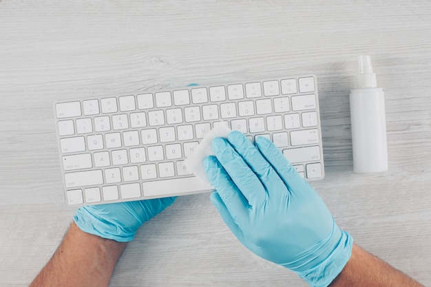 Ein Mann in Handschuhen, der eine Tastatur im hellen hölzernen Hintergrund mit Desinfektionsmittel desinfiziert.