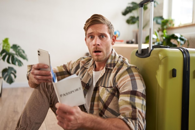 Kostenloses Foto ein mann mit einem verängstigten gesicht sitzt auf dem boden in der nähe eines koffers und hält ein smartphone und einen reisepass