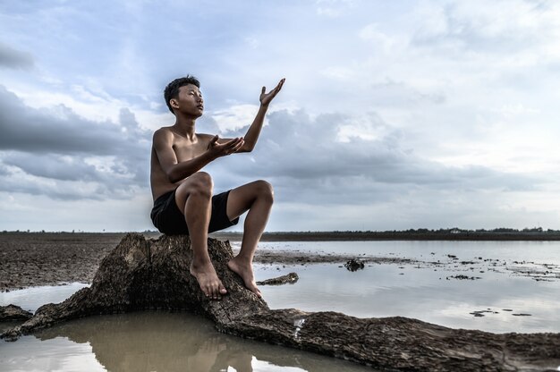 Ein Mann saß mit gebeugten Knien, machte ein Handsymbol, um am Baumstamm nach Regen zu fragen, und war von Wasser umgeben.