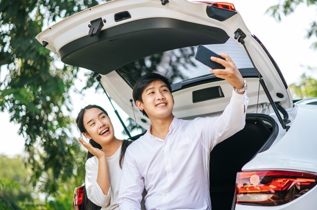 Ein Mann und eine Frau sitzen glücklich im Kofferraum eines Autos und machen ein Selfie am Telefon.