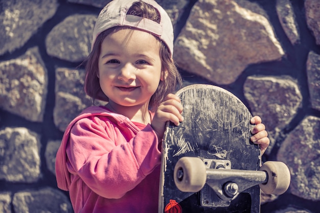 Ein modisches kleines Mädchen hält ein Skateboard und spielt draußen die schönen Gefühle eines Kindes.