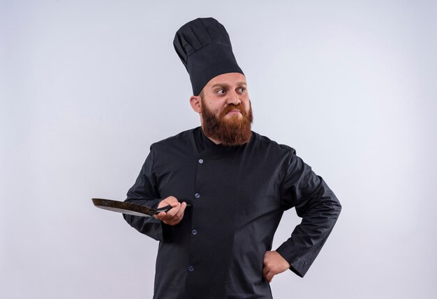 Ein nachdenklicher bärtiger Kochmann in der schwarzen Uniform, die schwarze Pfanne hält und Seite mit Händen auf Taille auf einer weißen Wand schaut
