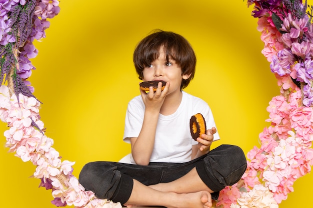 Ein niedlicher Junge der Vorderansicht, der Donuts um Blumen auf dem gelben Schreibtisch isst