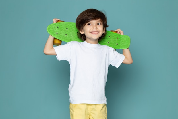 Ein niedlicher Junge der Vorderansicht im weißen T-Shirt und in den gelben Jeans, die grünes Skateboard auf dem blauen Raum halten
